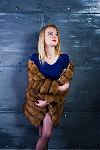 Elegante blonde girl wear on fur coat and combi dress posed against steel wall on studio