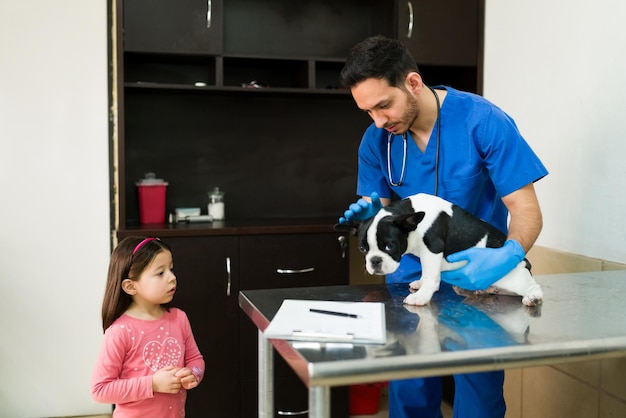 Elementary girl taking her beautiful pet to the vet because of an injury. Professional latin veterinarian working with a cute boston terrier at the exam room