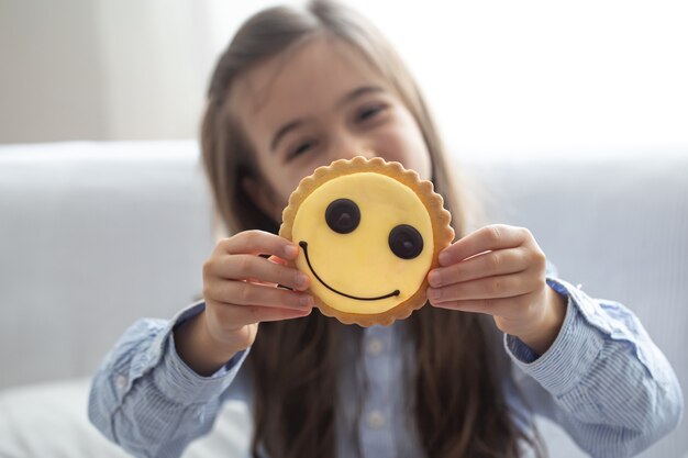 An elementary school girl in a shirt holds a bright yellow smiley cookie on a blurred background.