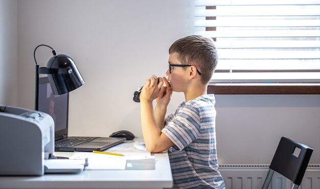 An elementary school student sits at a desk in front of a laptop and communicates via video link online at home.