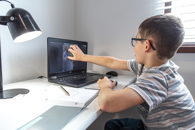 An elementary school student sits at a desk in front of a laptop and communicates with a friend via video link online, at home.