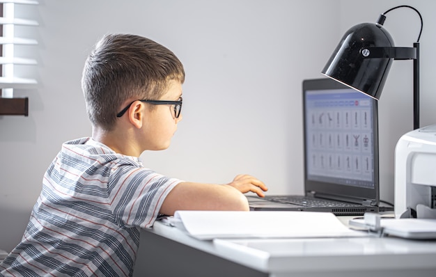 Free photo an elementary school student with glasses sits at a table with a laptop, does his homework online.