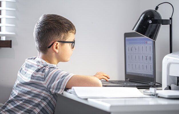 An elementary school student with glasses sits at a table with a laptop, does his homework online.