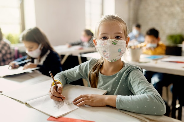 Elementary student with protective face mask learning in the classroom and looking at camera