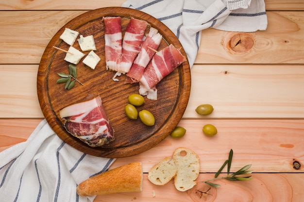An elevated view of bacon; olives; cheese and bread slices on wooden circular board over the table