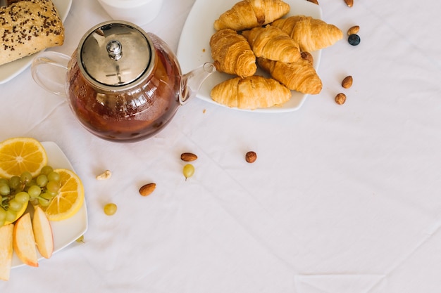 Free photo an elevated view of baked croissant; fruits; tea and dryfruits on white tablecloth