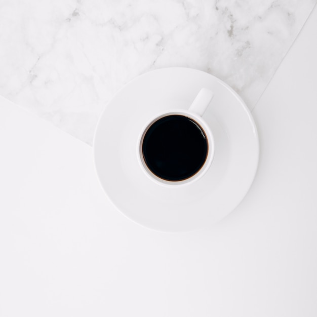 An elevated view of black coffee cup on saucer over the marble and white background