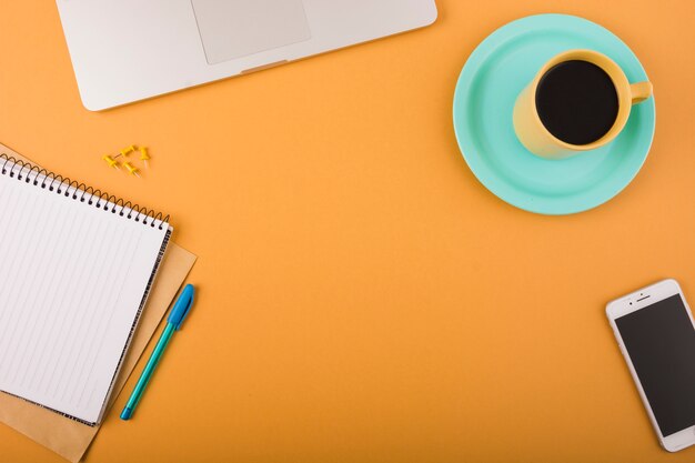 Elevated view of black coffee; smartphone; pen; pushpins; laptop and notepad on orange backdrop