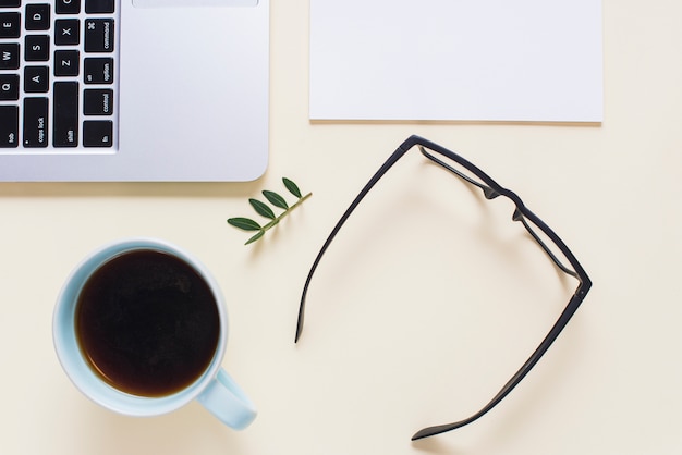 An elevated view of black eyeglasses; tea cup; laptop and paper on beige backdrop