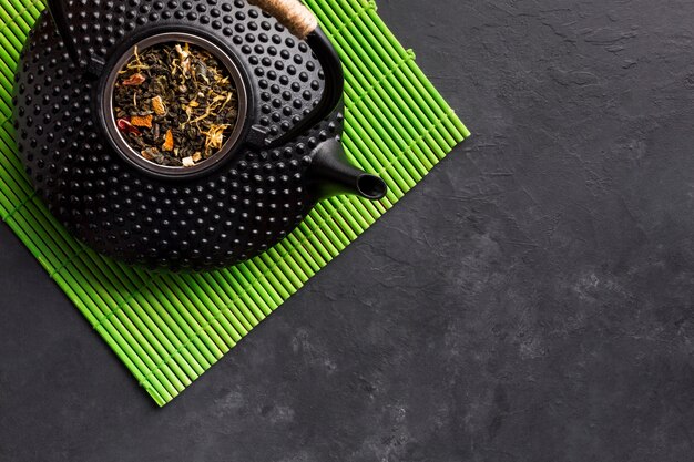 Elevated view of black teapot with dried tea herb on green placemat