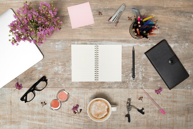 Elevated view of blank book surrounded by office supplies; make-up products; plant and laptop on old table