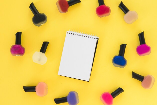 Elevated view of blank notepad surrounded by multi colored nail varnish bottle over yellow background