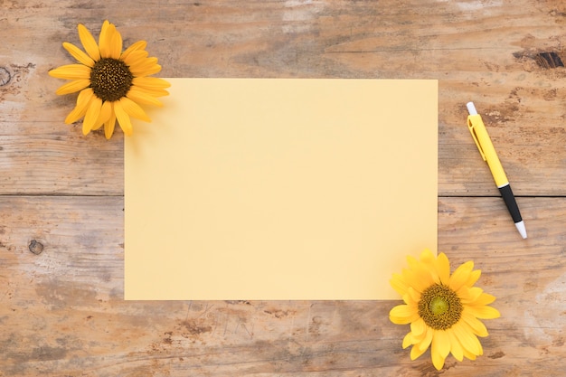 Elevated view of blank paper with yellow sunflowers and pen on wooden backdrop