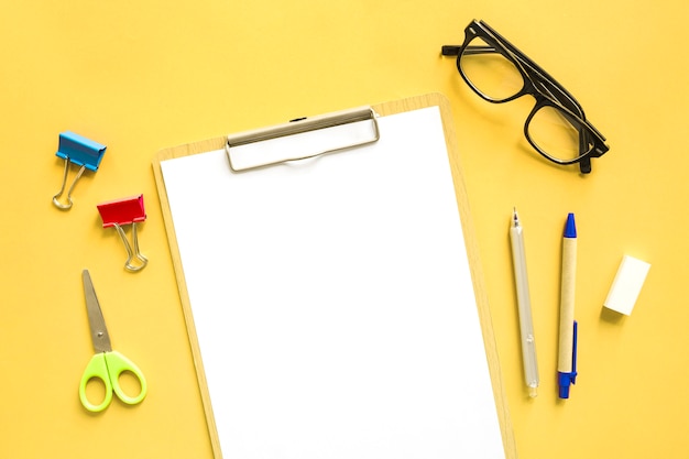 Elevated view of blank white paper near stationeries on yellow background