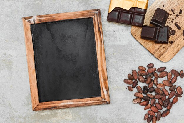 Elevated view of blank wooden slate, dark chocolate bar and cocoa beans on concrete background