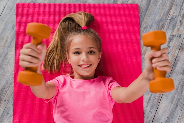 Free photo an elevated view of a blonde girl lying on pink carpet exercising with dumbbell