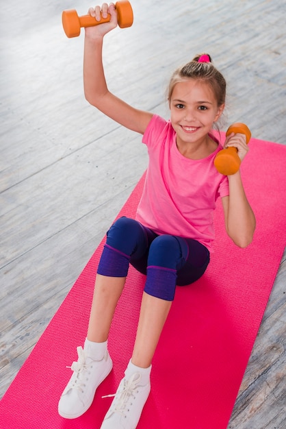 Free photo an elevated view of a blonde girl sitting on pink carpet exercising with dumbbell