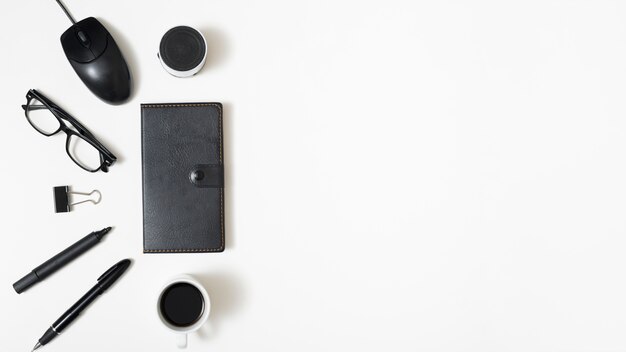 Elevated view of bluetooth speaker; mouse; eyeglass; paper clip; pen and coffee cup with diary on white background