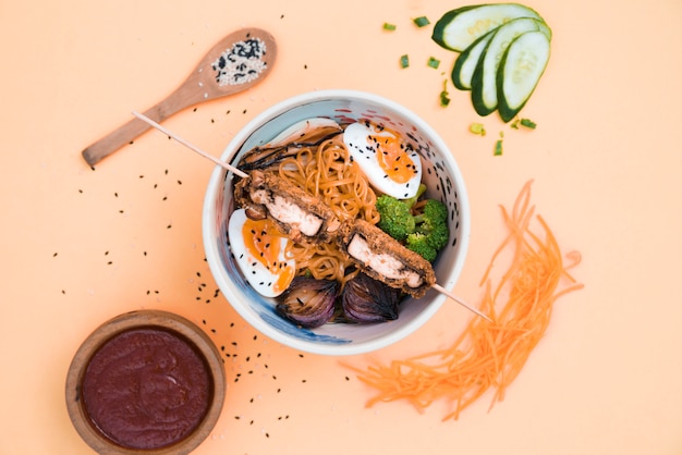 Free photo an elevated view of bowls with noodles; sauce; cucumber; carrot and sesame seeds on colored backdrop