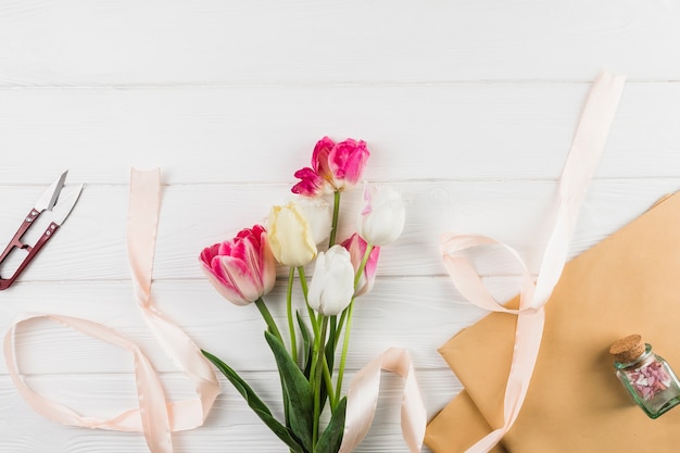 Free photo elevated view of brown paper; tulip flowers; ribbon and cutter against white desk