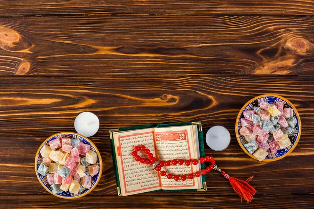 An elevated view of candles; red prayer beads with holy kuran and lukum bowls on wooden table