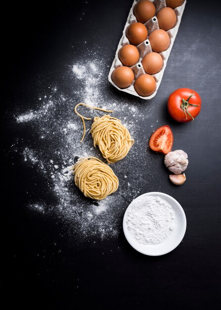 卵のカートンとカペッリーニパスタの立面図。ジューシートマト。にんにくと小麦粉のボウル