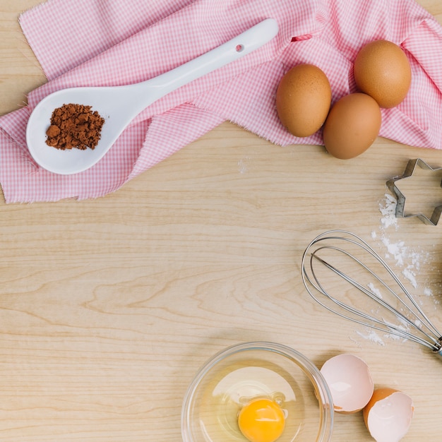 An elevated view of chocolate powder; eggs; egg yolk; whisk; and pastry cutter on wooden surface