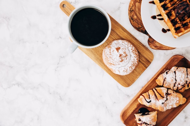 An elevated view of coffee cup; baked buns; croissant and waffles on wooden tray against marble textured background