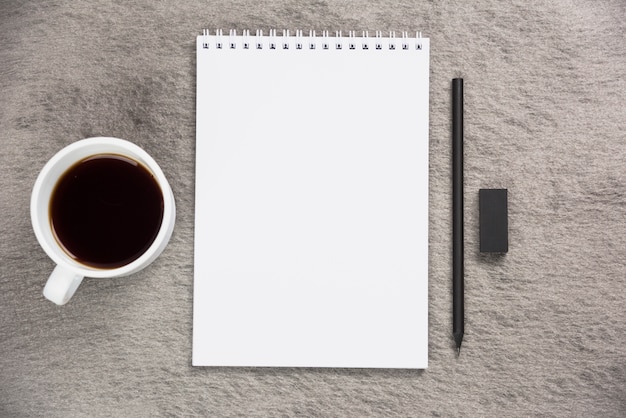 An elevated view of coffee cup; blank spiral notepad with black eraser and pencil on gray desk
