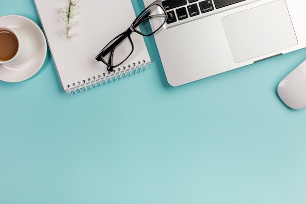 An elevated view of coffee cup,spiral notepad,eyeglasses,laptop and mouse on blue backdrop