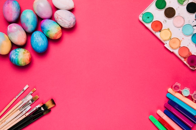 An elevated view of colorful easter eggs; brushes; felt tip pen and watercolor paint box on pink background