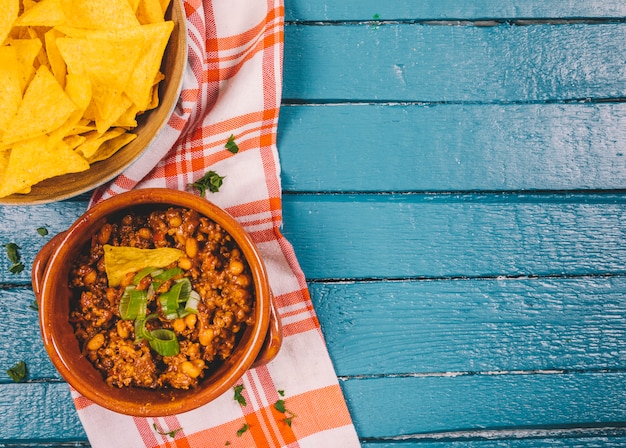 Elevated view of cooked ground beef in bowl with mexican nachos chips on blue wooden desk