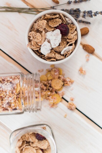 Elevated view of cornflakes in bowl and spilled jar of granola on wooden background