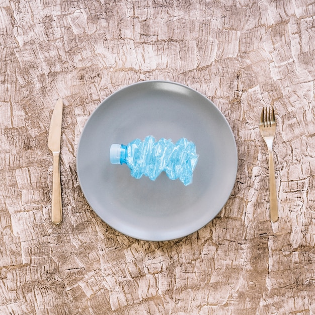 Free photo elevated view of crumpled plastic bottle on plate between fork and kitchen knife