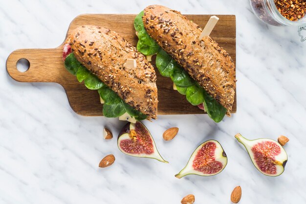 Elevated view of delicious hot dogs on wooden chopping board near fig slices; almonds and jar of chilli flakes over white marble