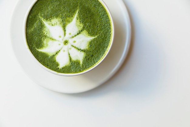 An elevated view of delicious matcha latte flower art cup on saucer over white background