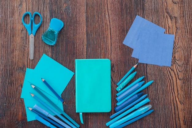 Elevated view of diary; drawing colors; scissor and colorful papers on textured wooden surface