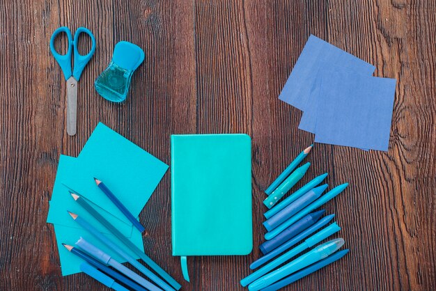 Elevated view of diary; drawing colors; scissor and colorful papers on textured wooden surface