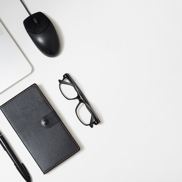 Elevated view of diary; eyeglass; pen; and laptop; mouse on white office table