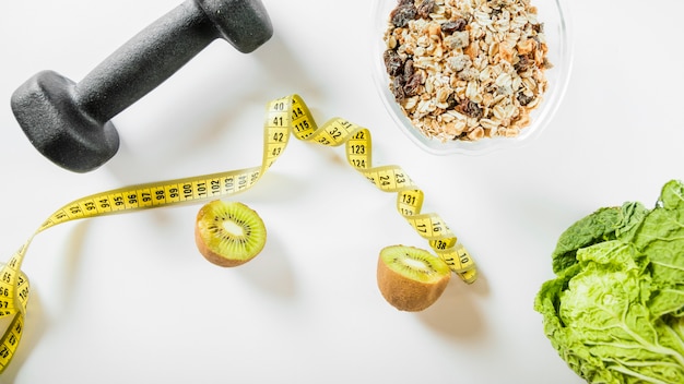 Elevated view of diet food with dumbbell and measuring tape on white backdrop
