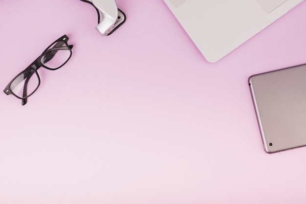 Elevated view of digital tablet; spectacles and laptop on pink backdrop