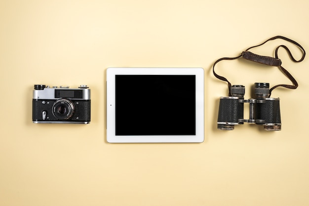 An elevated view of digital tablet with retro camera and binocular on beige background