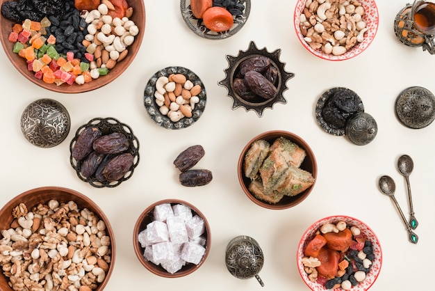 An elevated view of dried fruits; nuts; dates; lukum and baklava bowls over the white backdrop