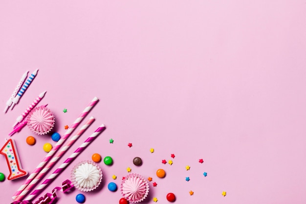 An elevated view of drinking straws; candles; gems; streamers and sprinkles on the corner of pink backdrop