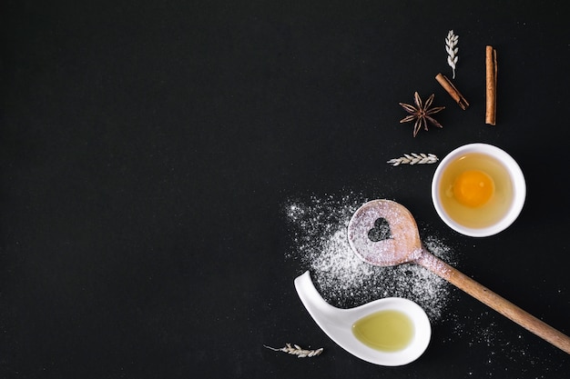 Free photo elevated view of egg; spices; grain; heart shape spoon; flour and oil on black surface