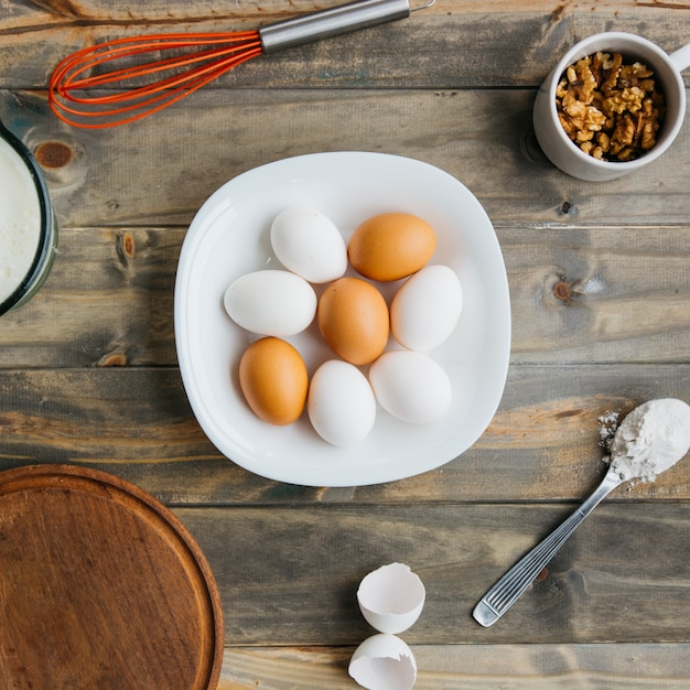 Elevated view of eggs; flour and walnut with whisk on wooden background