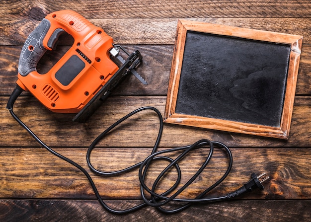 Elevated view of electric jigsaw and blank slate on wooden background