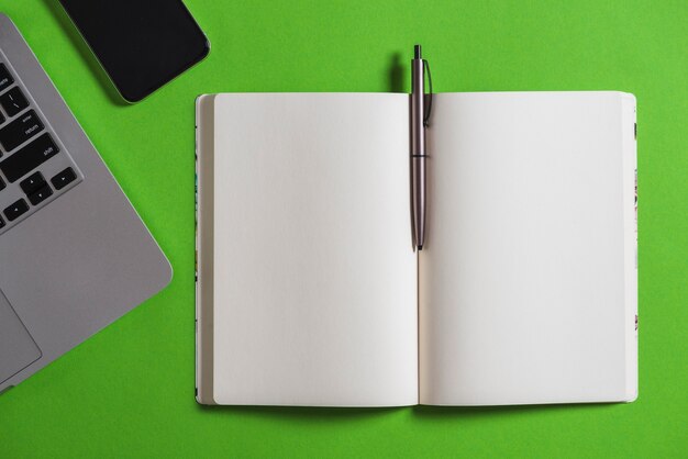 Elevated view of electronic gadget, notebook and pen on green background