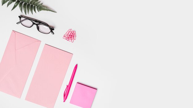 Elevated view of envelope; pen; adhesive note; artificial fern; spectacles and paper clips on white surface