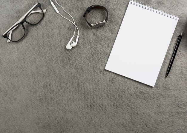 An elevated view of eyeglasses; earphone; wrist watch; spiral notepad and pen on gray desk